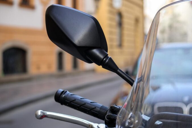 Gros plan sur un rétroviseur noir et le bord d'un parebrise moto transparent en ville.