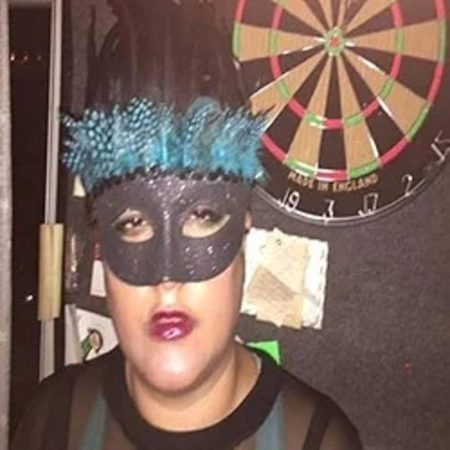 Dj Parisa portant un masque vénitien noir et une coiffe de plumes bleues devant une cible de fléchettes